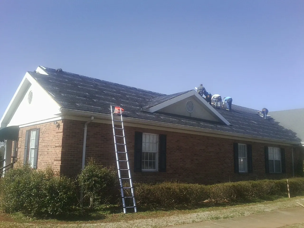 Crew tearing shingles off a brick ranch roof with a ladder set — Metal Roof Repair work by our crew in Dime Box