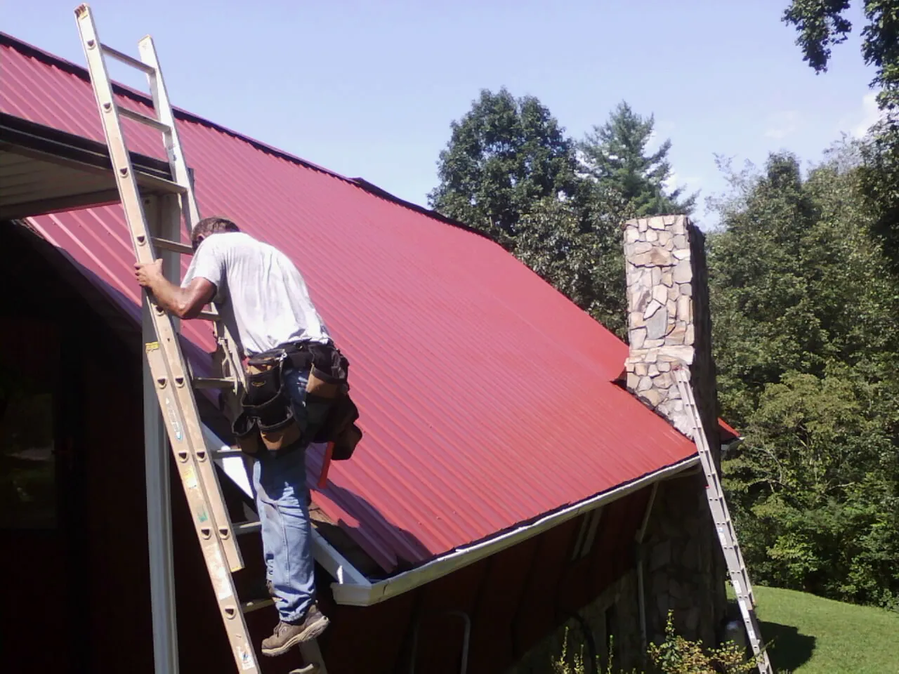 Roofer on a ladder beside a red metal roof and stone chimney — Soffit & Fascia Repair by Roofing Brothers in Dime Box